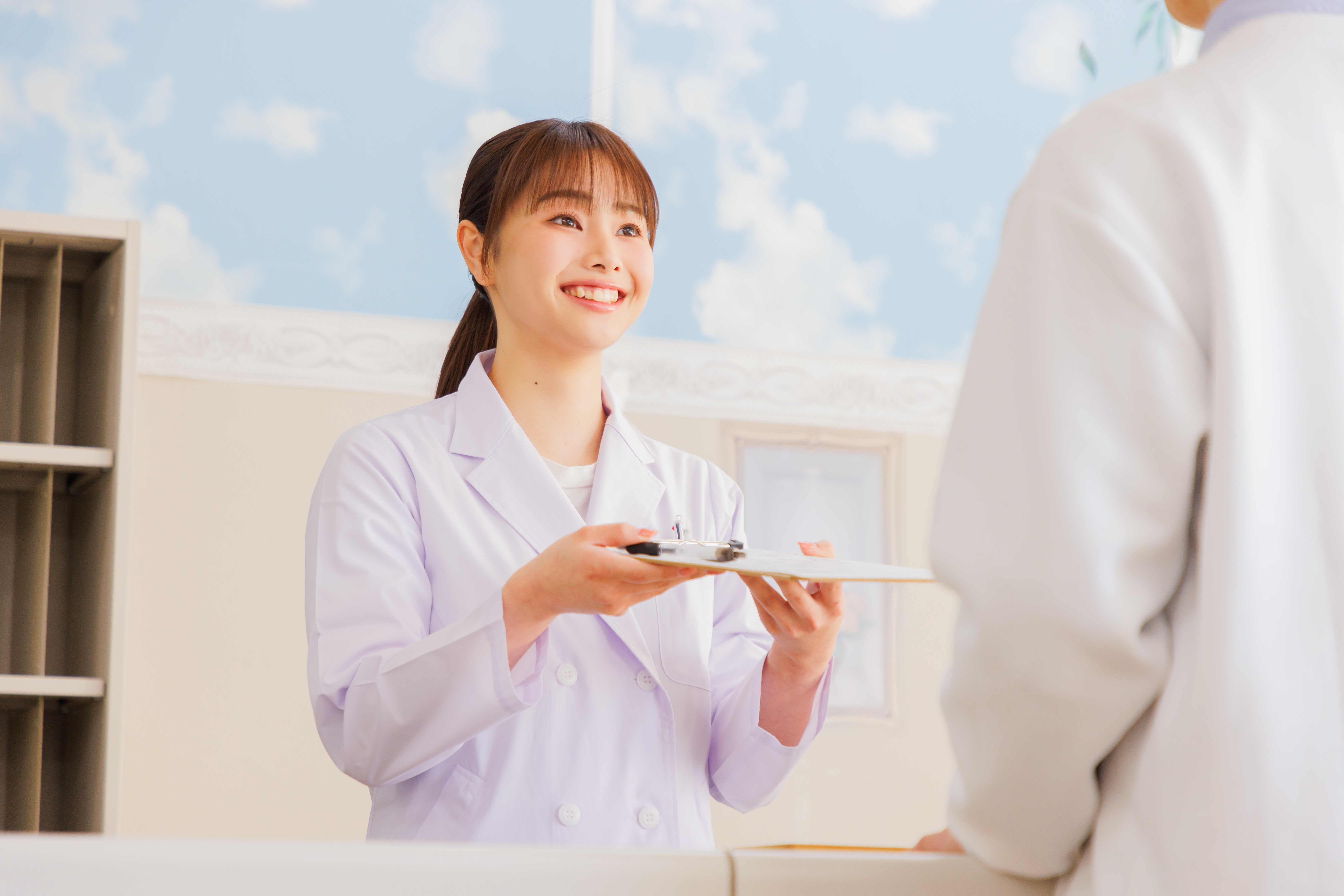 白衣を着た女性がカウンター越しの男性に、紙を挟んだバインダーを渡す様子