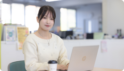 パソコンの前に座り、明るい表情でこちらを見つめる女子学生