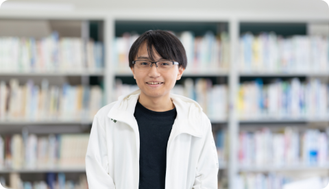 図書室の本棚を背に、眼鏡をかけて穏やかに微笑む男子学生